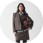 Eine Frau trägt einen braun karierten Blazer und einen dazu passenden Rock über einem gemusterten Pullover und einem Hemd mit Kragen und hält eine große braune Handtasche unter ihrem Arm. Sie steht vor einem schlichten weißen Hintergrund.
