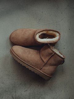Ein Paar braune, mit Shearling gefütterte Wildlederstiefel stehen auf einem grauen, strukturierten Boden, wobei ein Stiefel den anderen leicht überlappt.