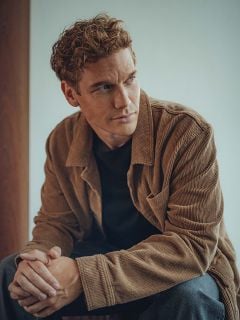 Ein Mann mit lockigem hellbraunem Haar sitzt in einem Haus und trägt eine braune Cordjacke über einem schwarzen Hemd. Er blickt nachdenklich zur Seite und hat die Hände locker verschränkt. Der Hintergrund ist sanft beleuchtet und neutral.