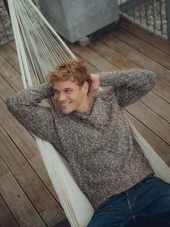 Ein junger Mann mit lockigem blondem Haar in einem braunen Pullover und Jeans entspannt sich in einer Hängematte, lächelt und blickt zur Seite. Die Hängematte steht auf einem Holzdeck mit einem Metallgeländer im Hintergrund.