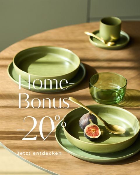 Ein runder Holztisch, gedeckt mit grünen Tellern, einer Tasse mit Untertasse, einem grünen Glas und goldenem Besteck. Auf einem Teller ruht eine halbierte Feige. Der Text lautet: "Home-Bonus -20%. Jetzt entdecken." Das Sonnenlicht wirft Schatten auf die Szene.