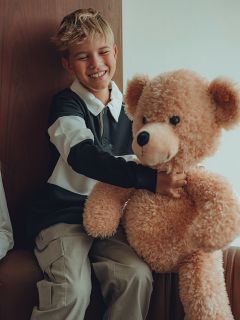 Ein lächelnder Junge mit blondem Haar sitzt in einem Haus und umarmt einen großen beigen Teddybär. Er trägt ein schwarz-weißes langärmeliges Hemd und eine helle Hose.