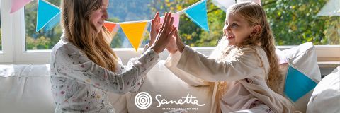 Zwei junge Mädchen sitzen im Schlafanzug auf einer Couch, lächeln und spielen ein Klatschspiel mit den Händen. Im Hintergrund hängen bunte Wimpel in der Nähe eines sonnenbeschienenen Fensters. Das Wort "Sanetta" erscheint unten in der Mitte.