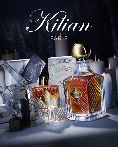 Eine Auslage mit Parfüms von Kilian Paris, darunter große und kleine Flaschen von "Angels' Share", umgeben von weißen Kerzen, einer dekorativen Schachtel, einem Band und einem Parfümetui, alles vor einem dunklen, eleganten Hintergrund.