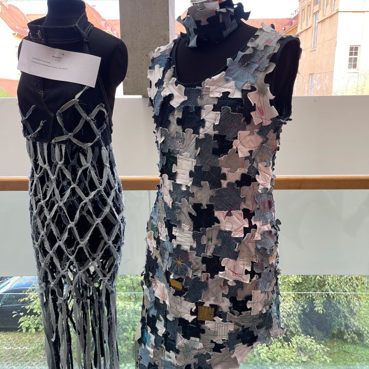 Zwei Schaufensterpuppen zeigen kreative Kleider; eine trägt ein netzartiges Outfit mit hängenden Strängen, die andere ein ärmelloses Kleid aus sich überlappenden bunten Puzzleteilen. Beide sind vor einem Fensterhintergrund ausgestellt.