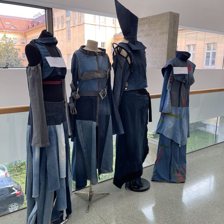 Vier Schaufensterpuppen zeigen avantgardistische Denim-Outfits mit geometrischen Formen und mehrschichtigen Details, darunter breite Gürtel, Riemen und ein dramatischer spitzer Hut. Sie stehen in Innenräumen vor großen Fenstern mit Blick auf historische Gebäude.
