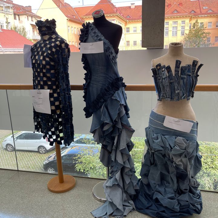 Drei Schaufensterpuppen zeigen kreative Kleidungsstücke aus upgecyceltem Denim mit gewebten, gerüschten und mehrlagigen Mustern. Sie stehen drinnen vor großen Fenstern mit einem Blick auf die Stadt im Hintergrund.