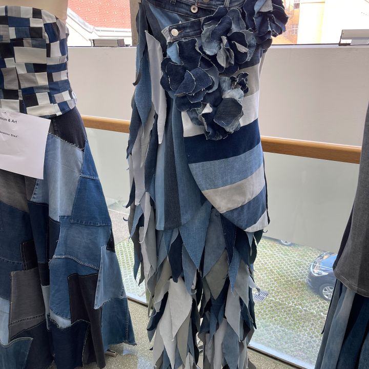 Ein auf einer Schaufensterpuppe ausgestelltes Kleid aus Jeansstoff in verschiedenen Farbtönen, der in blattähnlichen Lagen angeordnet und an der Taille mit Jeansblumen verziert ist. Daneben ist ein weiteres Kleid aus Denim-Patchwork teilweise sichtbar.