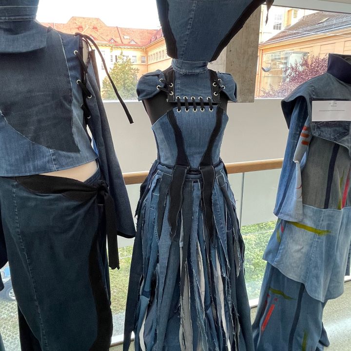 Ein Kleid aus verschiedenen Denim-Farbtönen mit geschnürtem Mieder, spitzen Schulterpartien und einem Rock aus langen Stoffstreifen, das auf einer Schaufensterpuppe in einem Raum neben anderen Denim-Kleidungsstücken ausgestellt ist.