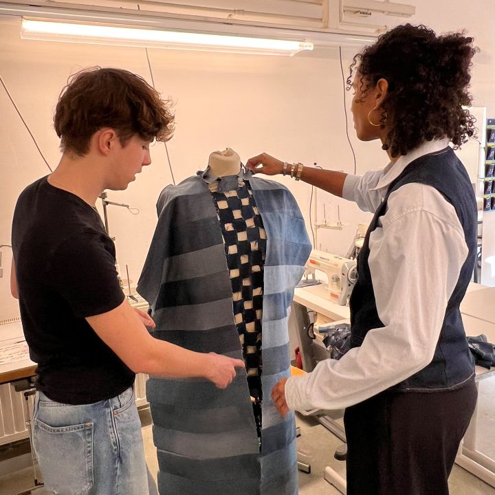 Zwei Personen in einem Modeatelier besprechen ein Kleidungsstück auf einer Kleiderform. Einer zeigt auf den Stoff, der blaue Streifen und ein kariertes Muster aufweist, während der andere das Kleidungsstück zurechtrückt. Im Hintergrund sind Nähmaschinen und Werkzeuge zu sehen.