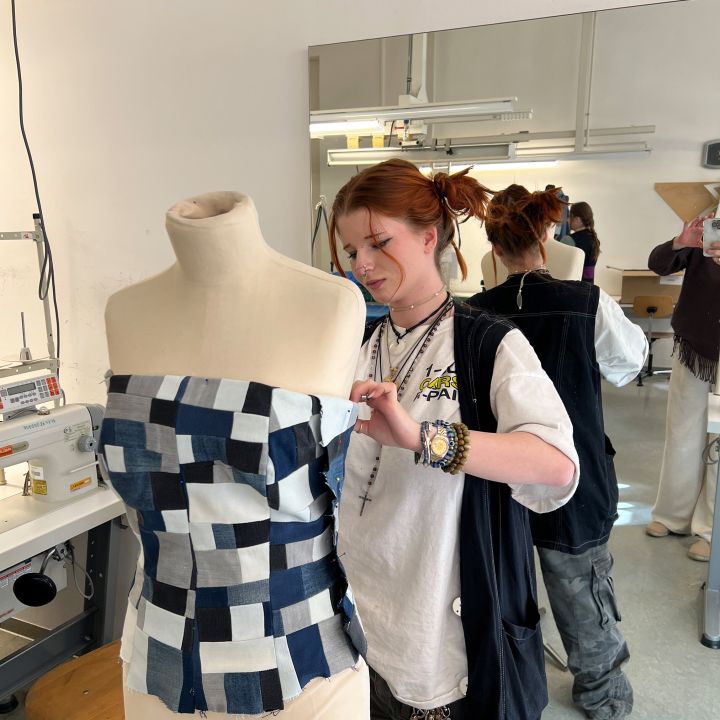 Eine Person heftet ein Patchwork-Jeansoberteil auf eine Kleiderform in einem Nähatelier, wobei im Hintergrund eine Nähmaschine, ein Spiegel und andere Personen zu sehen sind.