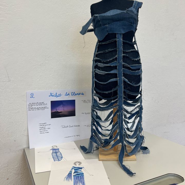 Eine Schaufensterpuppe zeigt ein Kleid aus übereinanderliegenden, zugeschnittenen Jeansstreifen, das einem Brustkorb ähnelt. Daneben befinden sich Skizzen von Modedesignern und eine Projekttafel mit dem Titel "Freiheit der Elemente" mit Text und dem Bild eines Sonnenuntergangs.