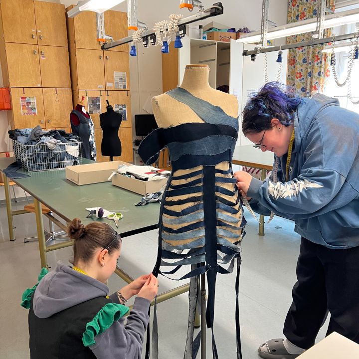 Zwei Personen arbeiten in einem Klassenzimmer gemeinsam an einer Kleiderform und bringen dunkle Stoffstreifen an, um ein einzigartiges Kleidungsstück zu entwerfen. Der Raum ist mit Arbeitstischen, Schaufensterpuppen, Holzschränken und bunten Vorhängen im Hintergrund ausgestattet.