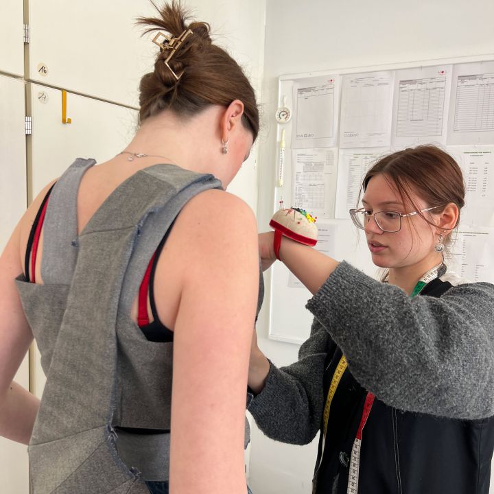 Zwei Frauen befinden sich in einer Schneiderei; eine trägt ein teilweise gefertigtes Kleidungsstück, während die andere es mit Nähwerkzeugen und einem Maßband um den Hals anpasst. An der Wand im Hintergrund sind Schnittmusterpapiere zu sehen.