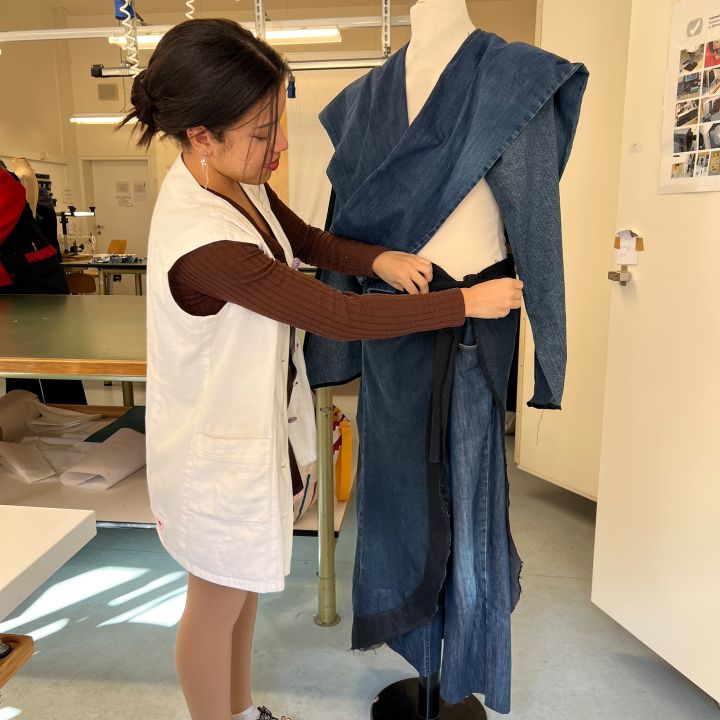 Eine Person, die ein braunes Oberteil, eine weiße Weste und Sandalen trägt, passt in einem hell erleuchteten Arbeitsbereich ein blaues kimonoähnliches Kleidungsstück auf einer Kleiderform an.
