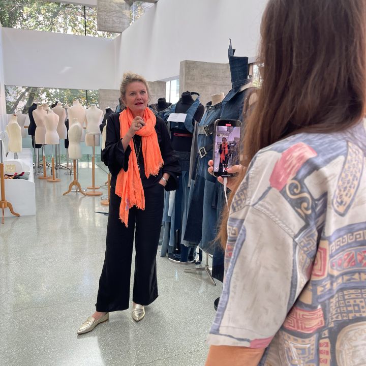 Eine Frau in schwarzem Outfit und leuchtend orangefarbenem Schal posiert zwischen Kleiderformen und Schaufensterpuppen, während eine andere Person sie mit einem Smartphone in einem hellen, modernen Studio fotografiert.