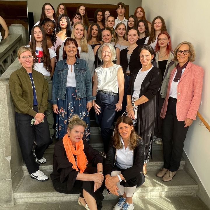 Eine Gruppe von Frauen und Mädchen posiert gemeinsam auf einer Treppe und lächelt in die Kamera. Einige stehen, andere sitzen auf der Treppe. Die Umgebung scheint ein Innenraum mit natürlichem Licht zu sein.