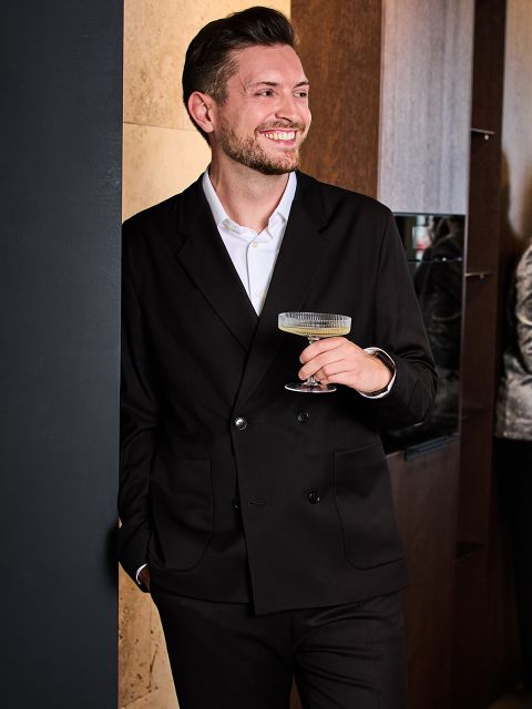Ein Mann in schwarzem Anzug und weißem Hemd hält lächelnd ein Cocktailglas in der Hand und lehnt lässig an einer Wand in einem Innenraum.