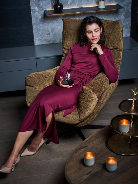 Eine Frau in einem langen burgunderroten Kleid sitzt auf einem braunen Sessel mit einem Glas Rotwein in der Hand, während auf einem Tisch in der Nähe Kerzen brennen.