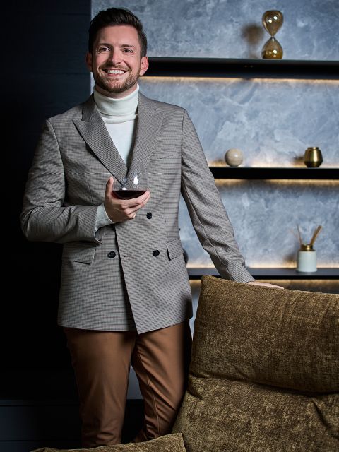Ein lächelnder Mann in einem karierten Blazer und einem weißen Rollkragenpullover hält ein Glas Wein in der Hand und steht neben einer braunen Couch. Im Hintergrund sind moderne Regale mit Dekorationsartikeln und sanfter Beleuchtung zu sehen.