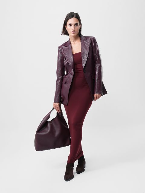 Eine Frau steht vor einem schlichten Hintergrund und trägt ein kastanienbraunes Kleid, einen passenden kastanienbraunen Lederblazer, braune Stiefeletten und eine große kastanienbraune Handtasche.