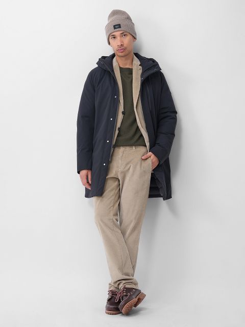 Ein Mann steht vor einer weißen Wand, trägt eine graue Mütze, einen dunklen Parka, einen olivgrünen Pullover, eine beigefarbene Hose und braune Schuhe, hat die Hände in den Taschen und einen Fuß über den anderen gekreuzt.