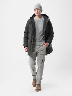 Ein Mann, der eine graue Mütze, eine dunkle Pufferjacke, einen hellgrauen Pullover, eine hellgraue Cargohose und braune Stiefel trägt, geht selbstbewusst vor einem schlichten weißen Hintergrund.