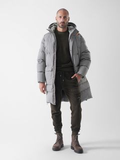 Ein Mann steht mit dem Gesicht nach vorne und trägt einen hellgrauen Kapuzenpullimantel, ein dunkles olivgrünes Sweatshirt und eine Jogginghose sowie braune Schnürstiefel vor einem schlichten weißen Hintergrund.