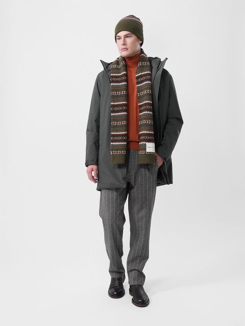 Eine Person steht vor einem schlichten Hintergrund und trägt einen grünen Mantel, einen orangefarbenen Rollkragenpullover, einen gemusterten Schal, eine grüne Strickmütze, eine graue Nadelstreifenhose und dunkle Schuhe.