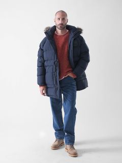Ein Mann steht vor einem schlichten weißen Hintergrund und trägt eine marineblaue Pufferjacke mit einer pelzgefütterten Kapuze, einen roten Pullover, blaue Jeans und hellbraune Schuhe.