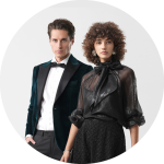 Ein Mann im Samt-Smoking und eine Frau in einer schwarzen Bluse und einem schwarzen Rock posieren gemeinsam vor einem hellgrauen Hintergrund.