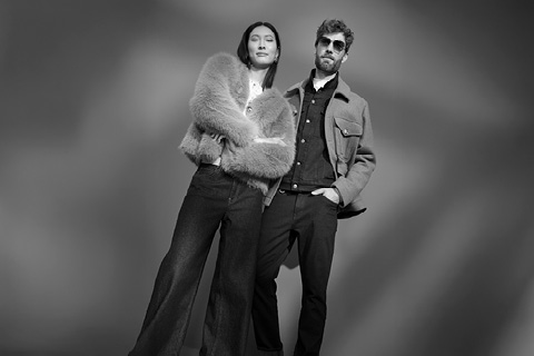 Zwei Personen posieren selbstbewusst in stilvollen Outfits; die eine trägt eine Pelzjacke, die andere eine Jacke und eine Sonnenbrille. Schwarz und weiß, Studiohintergrund.