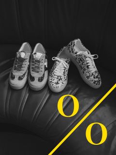 Ein Schwarz-Weiß-Foto von zwei Paar gemusterten Turnschuhen auf einem schwarzen Ledersitz. Eine gelbe diagonale Linie mit zwei gelben Buchstaben "O" ziert die untere rechte Ecke des Bildes.