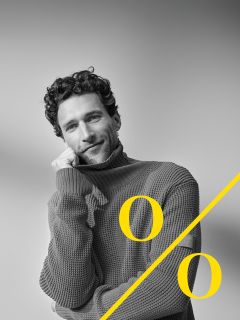 Ein Mann mit lockigem Haar und Bart trägt einen dicken Rollkragenpullover und stützt sein Gesicht lächelnd auf seine Hand. Ein großes gelbes Prozentzeichen ist diagonal über das Bild gelegt.