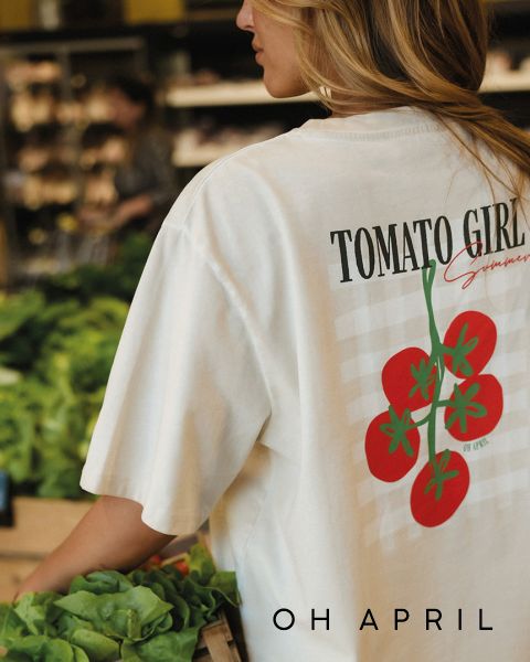 Eine Frau mit einem "Tomato Girl Summer"-T-Shirt steht neben frischem Salat in einem Lebensmittelgeschäft. Das "OH APRIL"-Logo erscheint am unteren Rand.