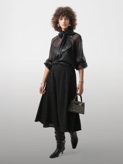 Eine Person, die eine durchsichtige schwarze Bluse mit Schleife, einen strukturierten schwarzen Midirock, schwarze Stiefel und eine kleine Nietenhandtasche trägt, steht vor einem schlichten Hintergrund.