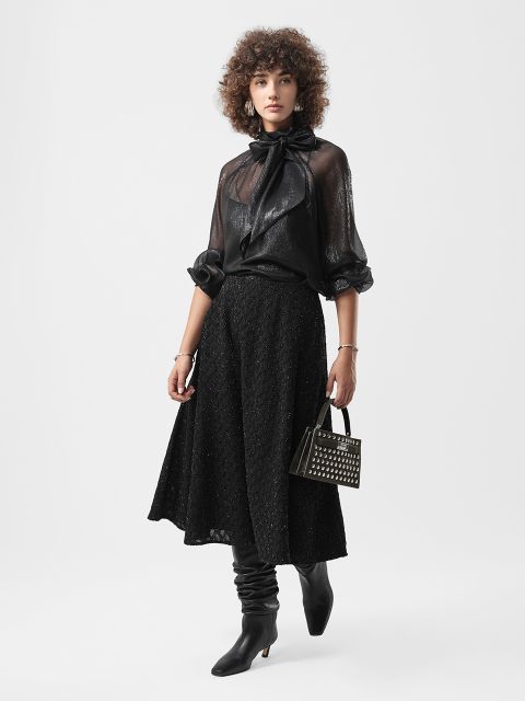 Frau in schwarzer, durchsichtiger Bluse mit Schleife, schwarzem Midirock, Stiefeln mit Absatz und einer kleinen, mit Nieten besetzten Handtasche vor einem schlichten Hintergrund.