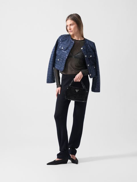 Eine Frau in einer strukturierten blauen Jacke, einer schwarzen Hose und schwarzen Flats hält eine schwarze Handtasche vor einem schlichten weißen Hintergrund.