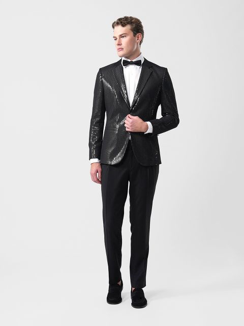 Ein Mann in einer schwarzen Pailletten-Smokingjacke, weißem Hemd, schwarzer Fliege und schwarzer Hose steht vor einem schlichten weißen Hintergrund.