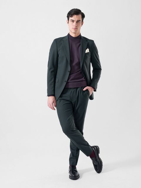 Ein Mann in einem dunkelgrünen Anzug mit weißem Einstecktuch, einem kastanienbraunen Pullover und schwarzen Schuhen steht selbstbewusst vor einem schlichten weißen Hintergrund.