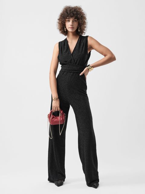 Frau mit lockigem Haar trägt einen ärmellosen schwarzen Overall, hält eine kleine rote Handtasche und steht vor einem schlichten weißen Hintergrund.