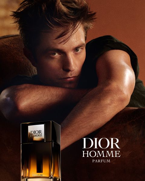Ein Mann lehnt auf einer braunen Fläche neben einem Flakon von Dior Homme Parfum, neben ihm steht der Text "DIOR HOMME PARFUM".