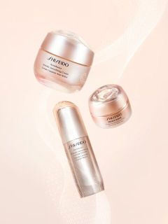 Drei Hautpflegeprodukte von Shiseido in eleganten, metallisch-rosa Behältern sind vor einem sanften, pfirsichfarbenen Hintergrund mit zarten, hellen Wirbelmustern zu sehen.