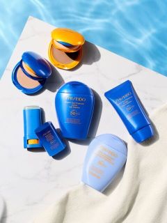 Verschiedene blaue und gelbe Sonnenschutzprodukte von Shiseido, die auf einer Marmorfläche am Pool angeordnet sind; ein beiges Handtuch ist teilweise zu sehen.