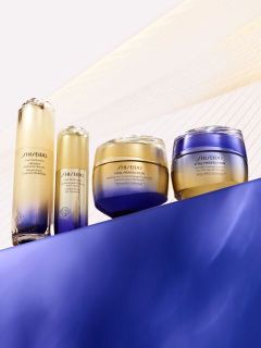 Vier Hautpflegeprodukte der Serie „Shiseido Vital Perfection“ in goldenen und blauen Flaschen und Tiegeln, angeordnet auf einer blauen Fläche vor einem hellen Hintergrund.