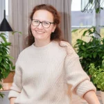 Eine Frau mit Brille und einem cremefarbenen Pullover lächelt, während sie in einem Innenraum in der Nähe von Grünpflanzen und großen Fenstern steht.