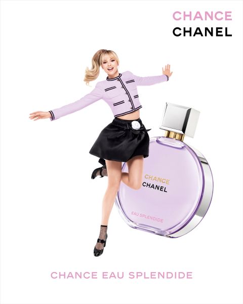 Eine lächelnde Frau in rosafarbener Jacke und schwarzem Rock springt neben einer großen Flasche des Parfums CHANEL Chance Eau Splendide auf weißem Hintergrund.