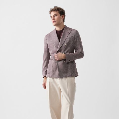 Eine Person, die einen lilafarbenen Zweireiher, ein kastanienbraunes Hemd und eine cremefarbene Hose trägt, steht vor einem schlichten weißen Hintergrund.