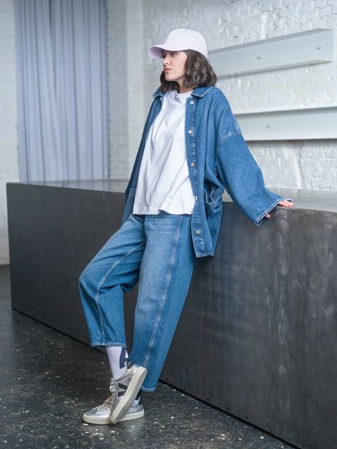 Eine Person, die eine Jeansjacke, ein weißes T-Shirt und eine weiße Mütze trägt, lehnt an einem dunklen Tresen in einem modernen Innenraum.