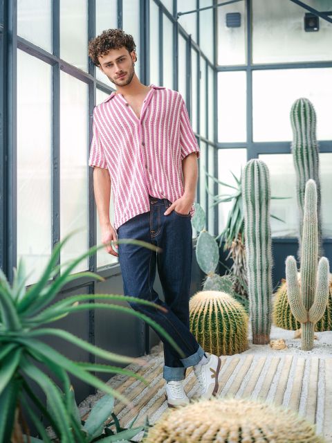 Ein Mann in einem rosa-weiß gestreiften Hemd und Jeans steht in einem Gewächshaus, umgeben von Kakteen und Sukkulenten, und schaut in die Kamera.
